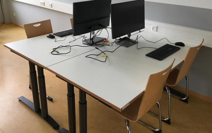 ine Tisch mit zwei PC-Bildschirmen, Kabeln, Tastaturen und stühlen davor