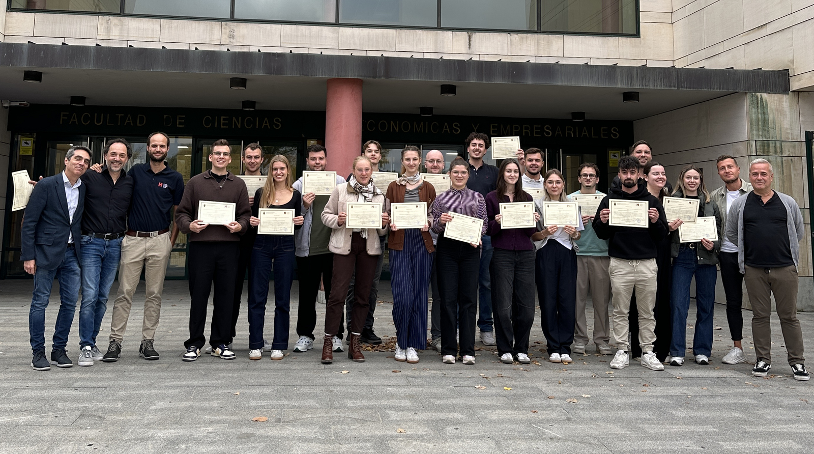 Eine Gruppe junger Menschen steht vor einem Universitätsgebäude und präsentiert ihre Zertifikate 