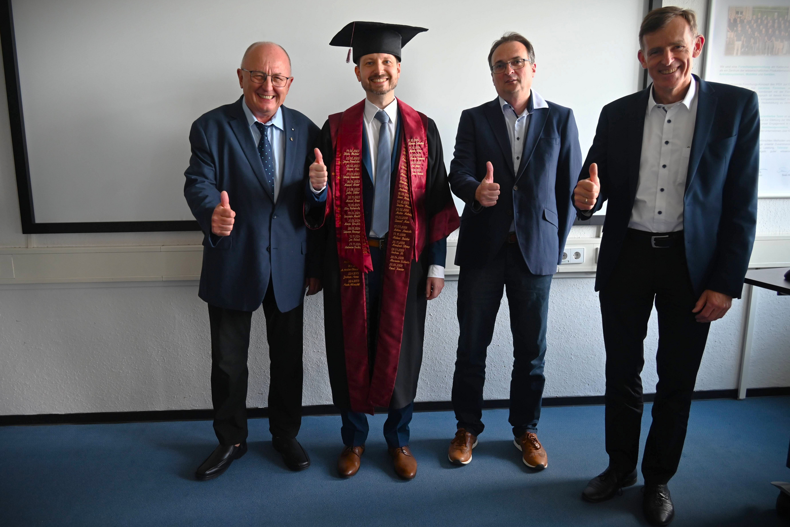 vier Männer stehen nebeneinander und strecken ihre Daumen in die Höhe, der zweite von rechts hat einen Doktorhut auf und eine Robe an